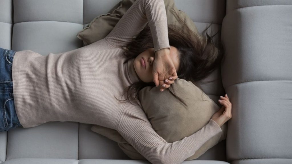 A tired woman lying on a couch with her arm covering her eyes, appearing emotionally and physically exhausted.